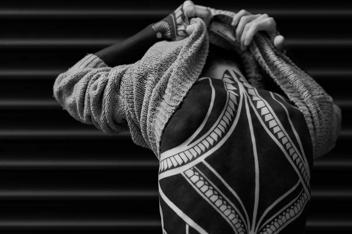 Man pulling off his jumper to show heavy blackwork tattoos on his back and arms