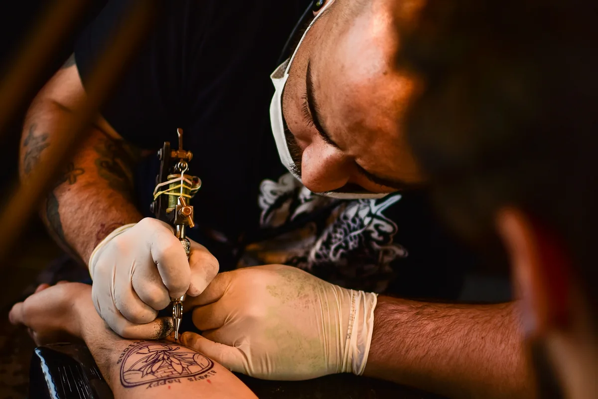 Masked tattooist concentrating on fine linework on a client's arm, one of the lower-pain spots on a tattoo pain chart